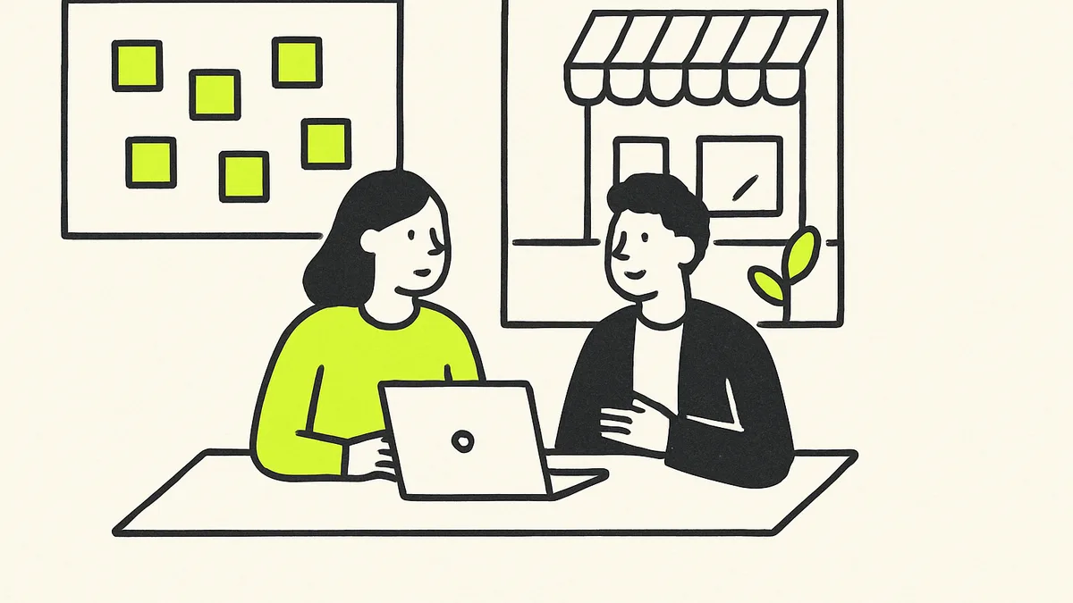 A local business advisor sitting across a table from a shop owner, both looking at a laptop screen with a workshop whiteboard behind them.